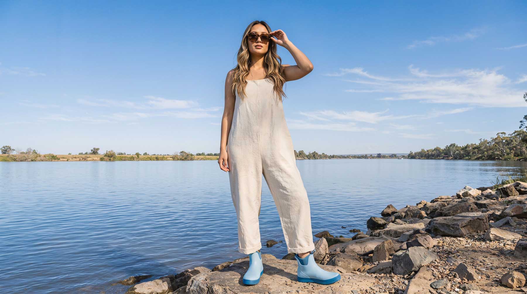 Woman in a white jumpsuit standing by a lake with sunglasses and gumboots on