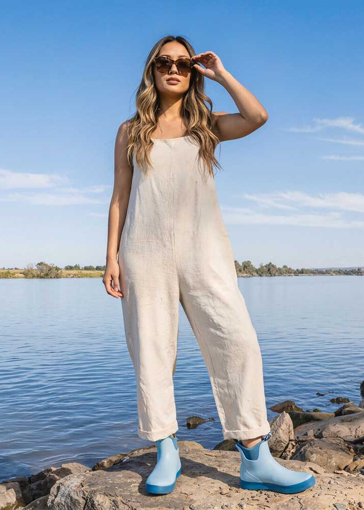 Woman in a white jumpsuit standing by a lake with sunglasses and gumboots on